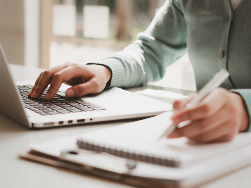 Personne travaillant à un bureau, utilisant un ordinateur portable d’une main tout en écrivant dans un carnet avec un stylo. La scène est lumineuse, avec une mise au point sur les mains, le clavier et le carnet de notes.