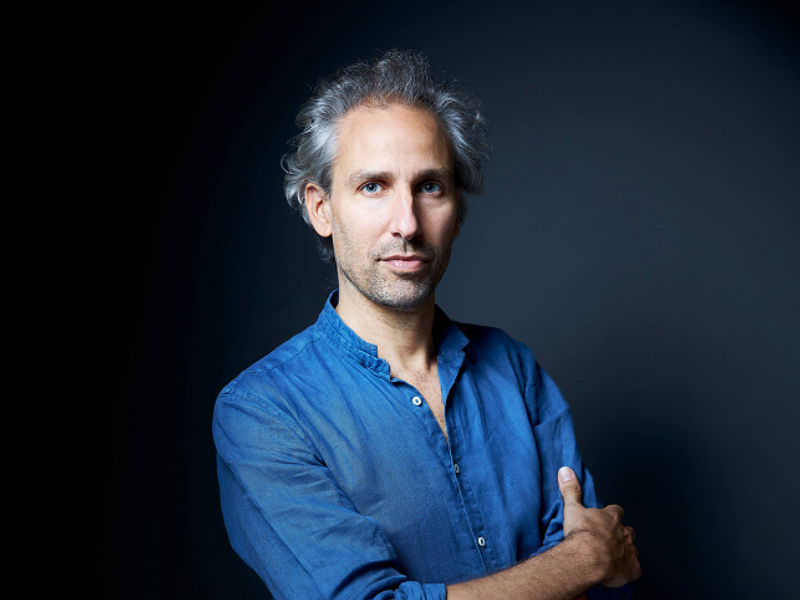 Portrait en studio d’une personne aux cheveux gris portant une chemise bleue à manches retroussées. La personne est photographiée de face, les bras croisés, sur un fond sombre aux nuances de bleu.