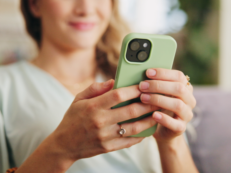 Photographie rapprochée montrant une personne tenant un smartphone vert dans ses mains. L'appareil mobile occupe le premier plan tandis que l'arrière-plan intérieur est volontairement flou pour mettre en valeur l'utilisation du téléphone.