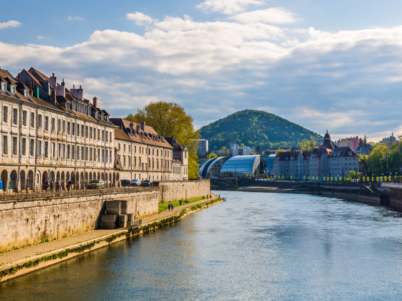 Vue urbaine le long d’une rivière, avec des bâtiments historiques, une colline verdoyante et un ciel nuageux.