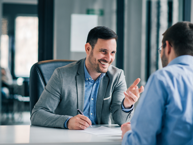 Personne en veste grise assise à un bureau dans un environnement de bureau moderne, tenant un stylo et échangeant avec une autre personne visible de dos. Des documents sont posés sur la table, avec un arrière-plan flou composé de baies vitrées et d'espaces de travail.