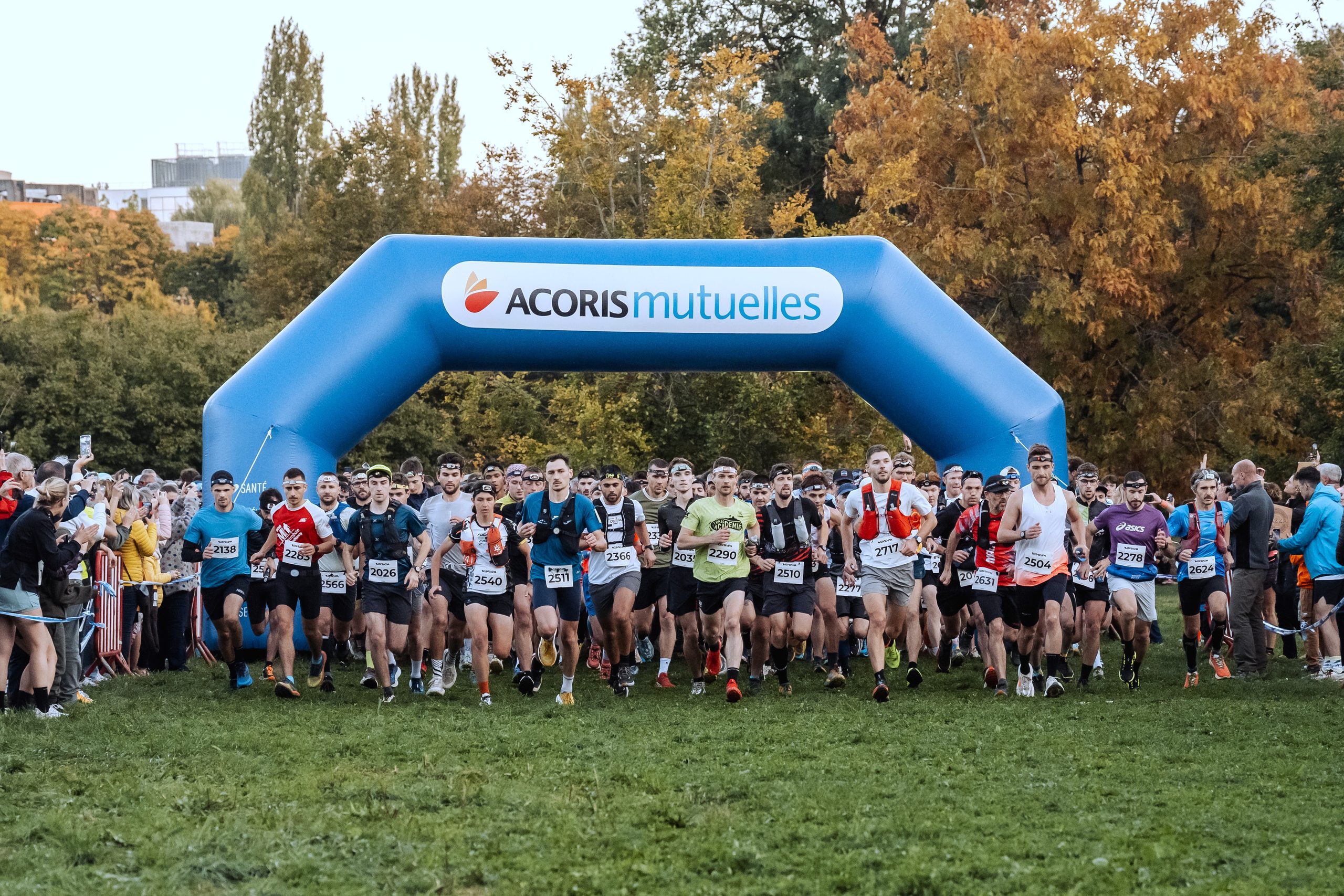 Départ d’une course nature dans un parc. Plusieurs dizaines de coureurs s’élancent sur une pelouse depuis une arche gonflable bleue portant le logo « ACORIS Mutuelles ». Des spectateurs sont rassemblés de chaque côté du parcours, tandis que des arbres aux couleurs automnales occupent l’arrière-plan.