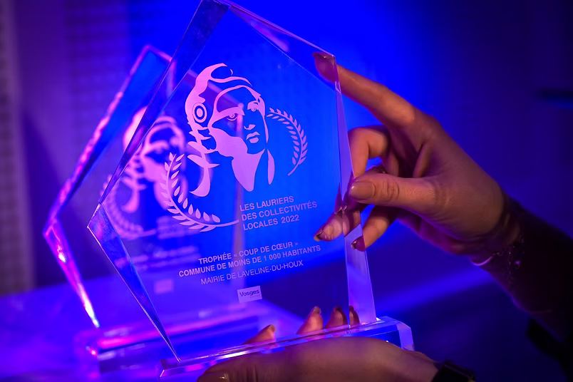 Gros plan sur un trophée transparent éclairé par des lumières bleues et violettes. Le trophée porte l’inscription « Les Lauriers des Collectivités Locales 2022 » et est tenu entre deux mains lors d’une cérémonie de remise de prix.