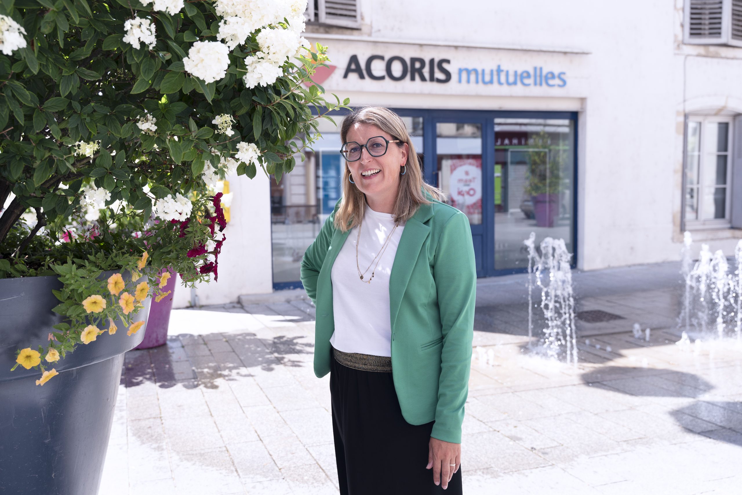 Personne portant une veste verte devant une agence Acoris Mutuelles, à proximité d’un massif de fleurs blanches et de jets d’eau sur une place urbaine.
