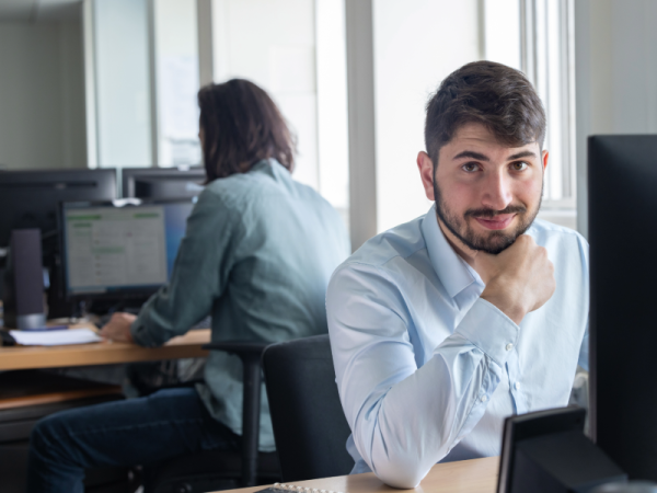 Personne assise à un poste de travail dans un bureau, regardant vers l’objectif, tandis qu’une autre personne travaille sur un ordinateur en arrière-plan.