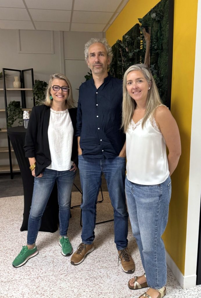 Trois personnes posent côte à côte dans un espace intérieur moderne. L'une porte une veste noire, un haut clair, un jean et des baskets vertes. La personne au centre porte une chemise bleu foncé et un jean. La troisième porte un débardeur blanc et un jean clair. À l'arrière-plan, on distingue un mur végétalisé, un mur jaune, des étagères décoratives et quelques plantes.