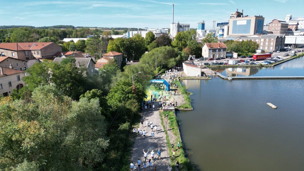 Vue aérienne de la Color Run longeant un canal dans un environnement urbain et verdoyant. Plusieurs dizaines de participants parcourent le chemin tandis qu’un nuage de poudre colorée marque un point d’animation près d’une arche bleue.