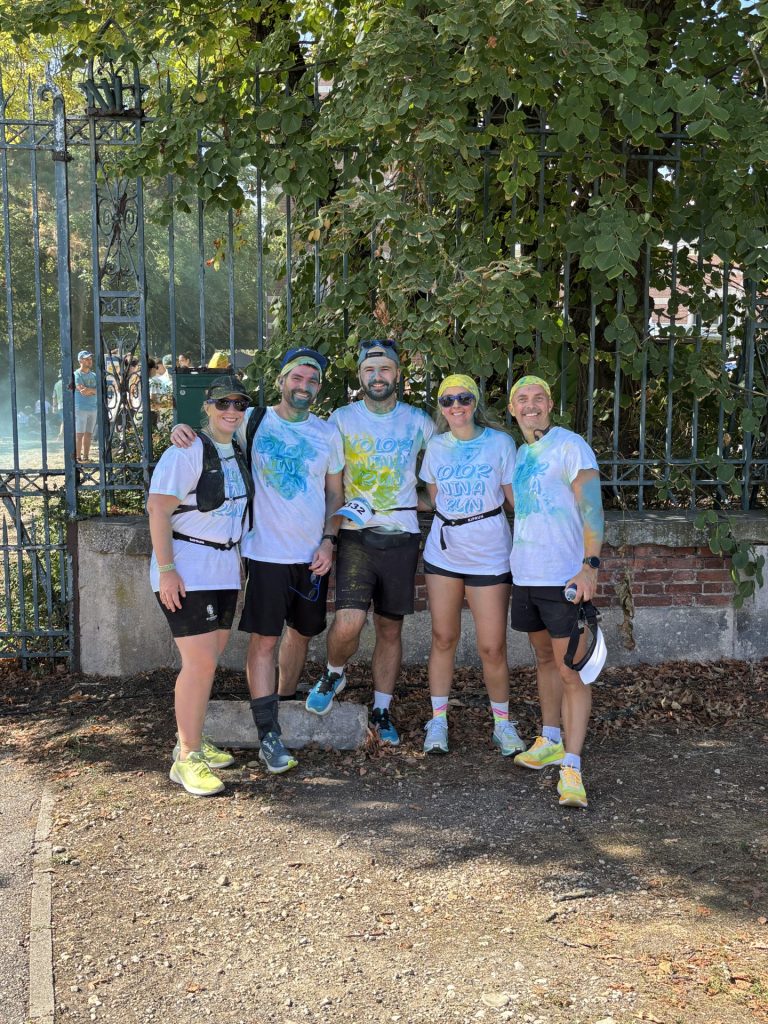 Cinq participants à la Color Run posent devant une grille et des arbres après la course. Les t-shirts blancs sont recouverts de poudre colorée bleue, jaune et verte, caractéristique des courses colorées, tandis que les participants portent encore leurs dossards et équipements de course.