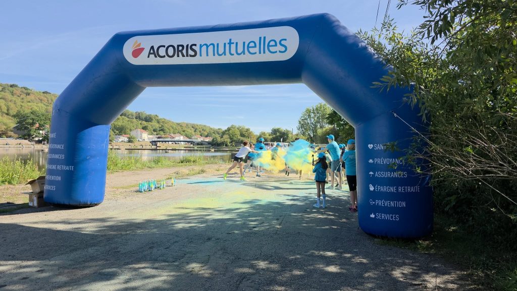 Des participants franchissent une arche gonflable bleue aux couleurs d’Acoris Mutuelles lors d’une Color Run organisée au bord d’un plan d’eau. Des bénévoles projettent des poudres colorées bleues et jaunes sur le parcours tandis que les coureurs avancent sous l’arche.