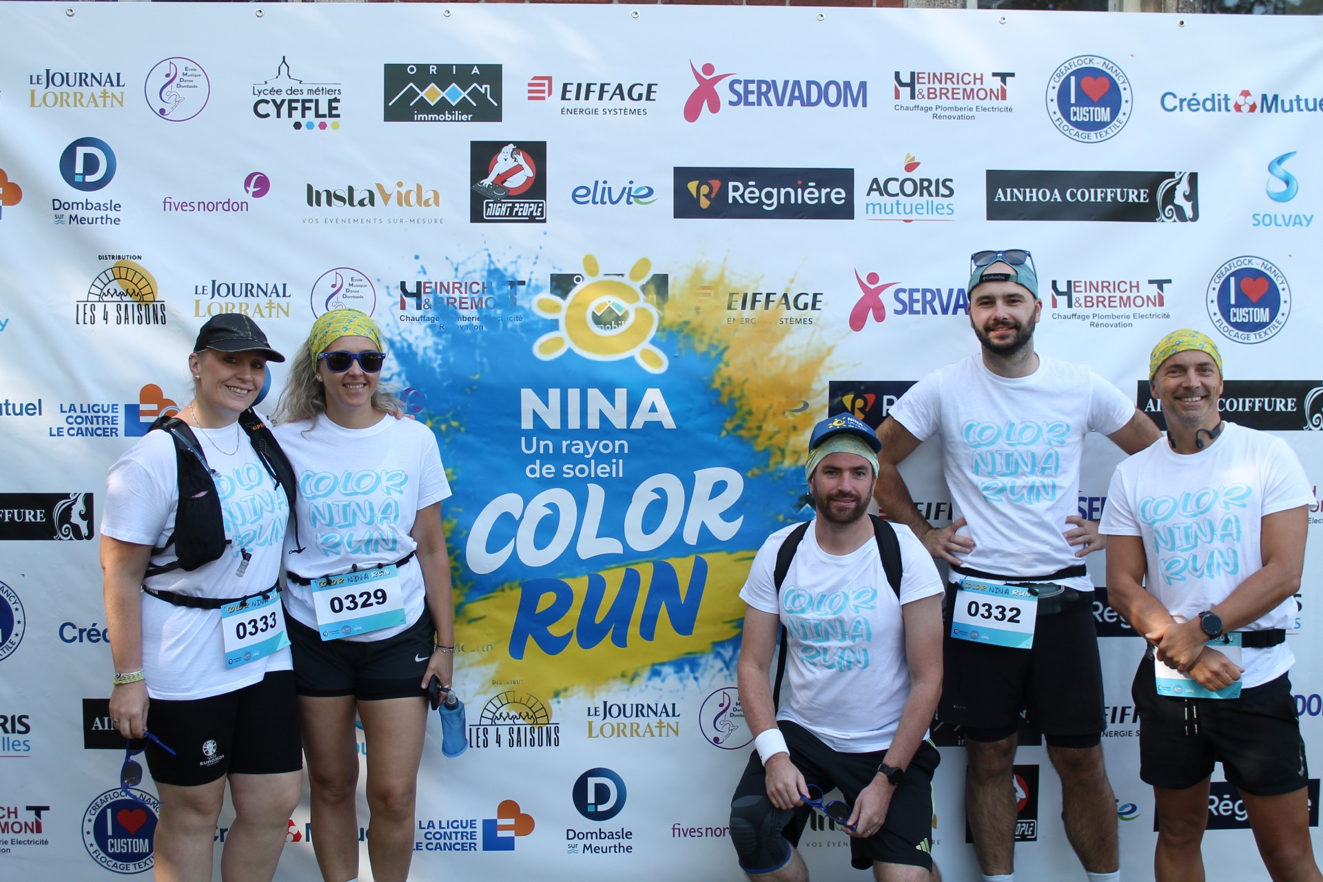 Cinq participants à la Color Run Nina posent devant un mur de partenaires et sponsors. Les participants portent des t-shirts blancs « Color Nina Run » et des dossards de course. Au centre, une grande affiche bleue et jaune affiche le message « NINA, un rayon de soleil - Color Run ».