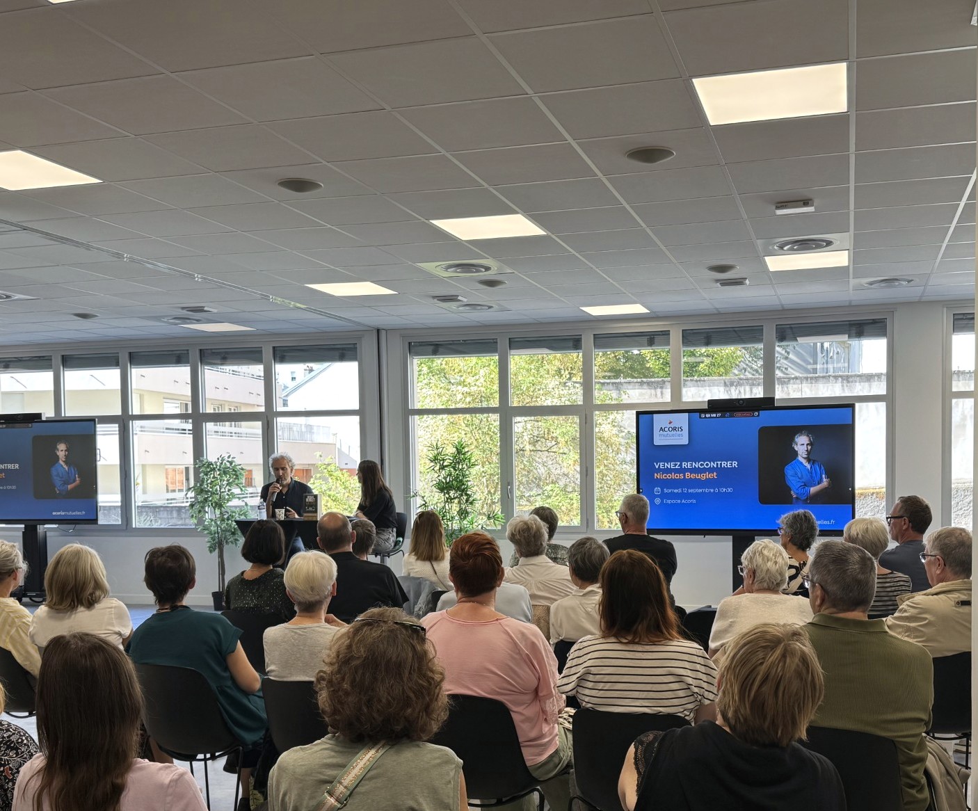 Une conférence se déroule dans une salle lumineuse devant un public assis. Deux personnes échangent sur scène devant de grands écrans affichant la promotion d'une rencontre organisée par Acoris Mutuelles avec Nicolas Beuglet. De larges baies vitrées laissent entrer la lumière naturelle, tandis que plusieurs dizaines de participants assistent à l'événement.