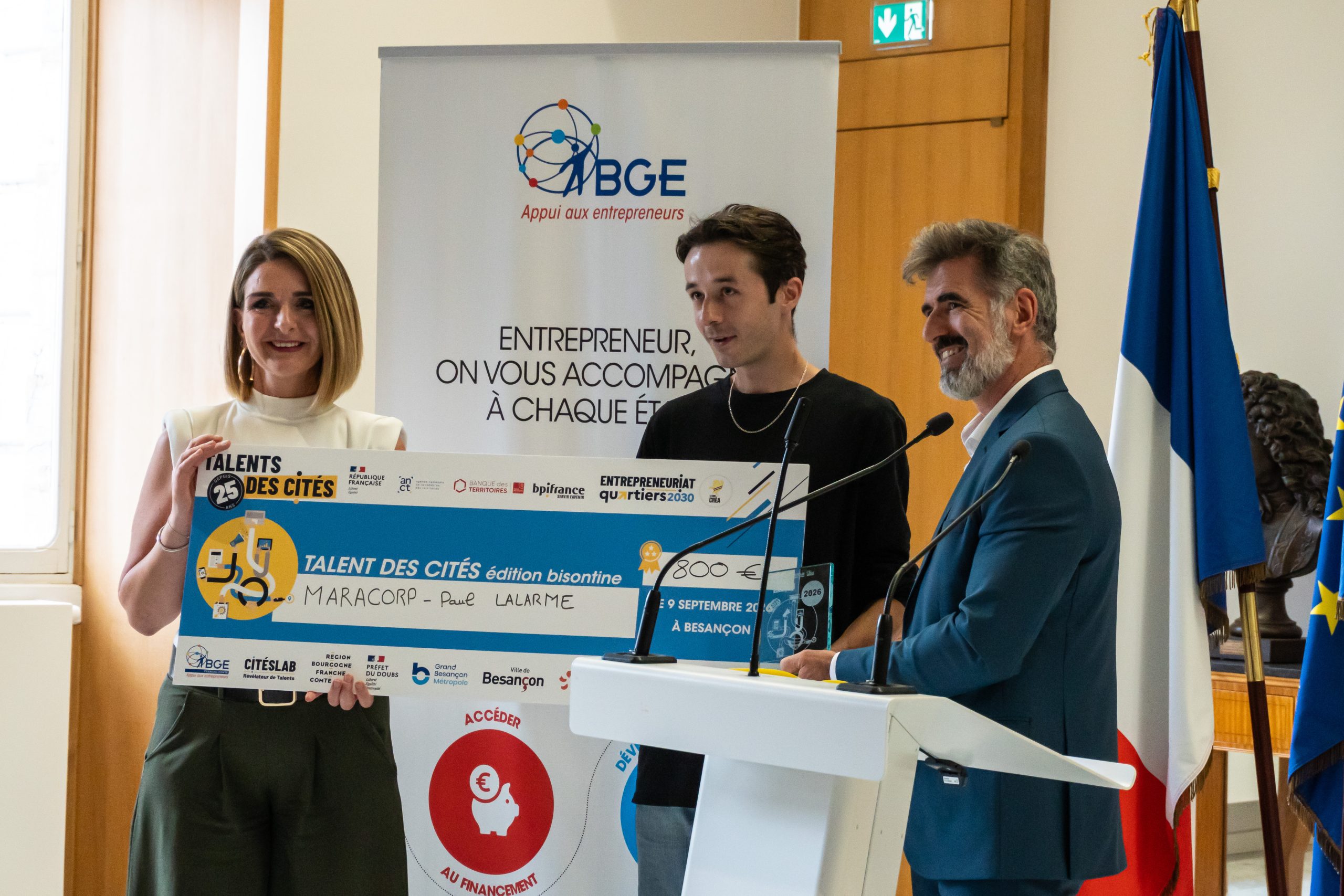Lors d’une cérémonie de remise de prix, trois personnes se tiennent devant un pupitre et présentent un chèque géant du concours « Talents des Cités ». À l’arrière-plan, une bannière de l’ABGE (Appui aux entrepreneurs) affiche le message « Entrepreneur, on vous accompagne à chaque étape ». Des drapeaux français et européens sont visibles sur le côté de la scène. Le chèque, attribué à « MARACORP - Paul Lalarme », indique une dotation de 800 € dans le cadre de l’édition bisontine de « Talents des Cités ».