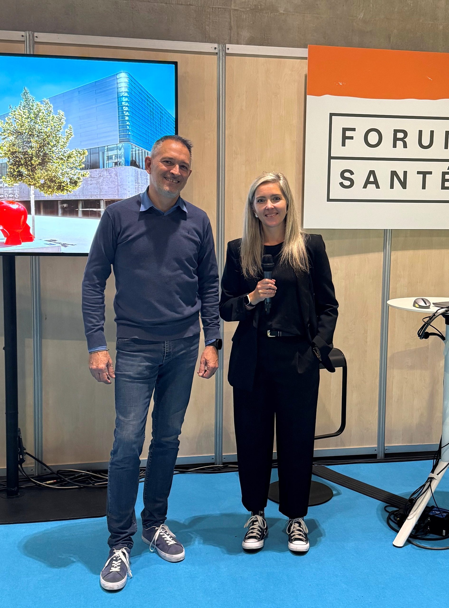 Deux personnes se tiennent sur un stand lors d'un évènement professionnel. L'une d'elles tient un micro. À l'arrière-plan, un écran affiche une photographie d'un bâtiment moderne et un panneau comporte l'inscription "Forum Santé". Cette image illustre un échange autour des enjeux de la santé, de la prévention et de la sensibilisation en milieu professionnel.